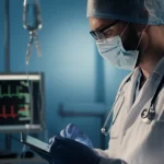 Medico in un pronto soccorso moderno che esamina i dati su un tablet, sullo sfondo un monitor con un tracciato cardiaco e una flebo. Atmosfera di urgenza controllata. Zoom lens, 50mm, depth of field, illuminazione clinica ma non fredda.