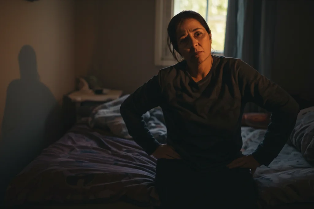 Fotografia ritratto, obiettivo 35mm, di una persona di mezza età seduta sul bordo del letto alle prime luci dell'alba, con espressione stanca e pensierosa, una mano appoggiata delicatamente sull'anca. Luce naturale soffusa dalla finestra. Profondità di campo con sfondo della camera leggermente sfocato. Palette di colori tenui.
