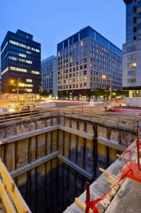 Fotografia grandangolare, 20mm, di uno scavo profondo per un parcheggio sotterraneo situato strategicamente tra due moderni edifici alberghieri in un contesto urbano denso. Messa a fuoco nitida su tutta la scena, lunga esposizione utilizzata per catturare leggere scie luminose del traffico circostante nelle strade adiacenti, suggerendo l'attività urbana che continua durante i lavori. Illuminazione crepuscolare controllata che bilancia le luci artificiali del cantiere con la luce ambientale.