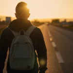 Immagine simbolica e fotorealistica: un giovane adulto cammina da solo su una strada al tramonto, voltato di spalle verso l'osservatore, dirigendosi verso un orizzonte incerto ma con una luce fioca all'orizzonte. Obiettivo grandangolare 24mm, luce calda del tramonto, leggera profondità di campo per mantenere a fuoco sia la figura che l'ambiente circostante, evocando il tema della transizione e delle sfide future.