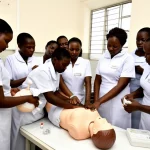 Fotografia molto realistica in stile reportage. Un gruppo di studentesse infermiere-ostetriche tanzaniane in divisa chiara è riunito attorno a un lettino da parto in un laboratorio di simulazione ben illuminato. Stanno praticando attivamente le procedure di gestione dell'emorragia post-partum su un manichino avanzato (MamaNatalie). Una studentessa applica un massaggio uterino mentre un'altra prepara farmaci. Un formatore osserva attentamente. Obiettivo prime 35mm per un'inquadratura ampia ma intima, luce naturale proveniente da una finestra laterale, colori vividi ma realistici, focus nitido sull'interazione centrale.