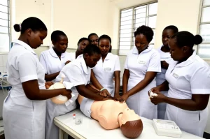 Fotografia molto realistica in stile reportage. Un gruppo di studentesse infermiere-ostetriche tanzaniane in divisa chiara è riunito attorno a un lettino da parto in un laboratorio di simulazione ben illuminato. Stanno praticando attivamente le procedure di gestione dell'emorragia post-partum su un manichino avanzato (MamaNatalie). Una studentessa applica un massaggio uterino mentre un'altra prepara farmaci. Un formatore osserva attentamente. Obiettivo prime 35mm per un'inquadratura ampia ma intima, luce naturale proveniente da una finestra laterale, colori vividi ma realistici, focus nitido sull'interazione centrale.