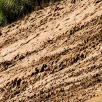 Immagine drammatica di una colata di fango in movimento lungo un pendio naturale, catturata con un teleobiettivo zoom (200mm) per comprimere la prospettiva; alta velocità dell'otturatore per congelare parzialmente il movimento del fango, mostrando schizzi e texture, luce naturale intensa che crea forti contrasti.