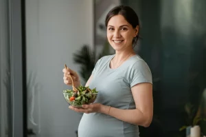 Donna incinta sorridente che tiene in mano una ciotola di insalata fresca con semi e noci, luce naturale morbida, ritratto 35mm, colori caldi e luminosi, profondità di campo.