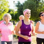 Gruppo di donne mature sorridenti in abbigliamento sportivo che camminano a passo svelto in un parco cittadino durante una giornata di sole, fotografia sportiva, obiettivo zoom 70-200mm, messa a fuoco sul gruppo in primo piano, sfondo leggermente sfocato, movimento catturato con velocità dell'otturatore media.