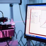 Immagine fotorealistica di un setup sperimentale per sensori a prisma LMR. Un prisma BK7 con un rivestimento sottile multistrato è illuminato da un raggio laser focalizzato (633nm), con la luce riflessa catturata da un fotorilevatore. Accanto, uno schermo di computer mostra l'interfaccia grafica del simulatore Sim-LMR con curve di riflettanza LMR ben definite. Stile high-tech, obiettivo 50mm prime, profondità di campo ridotta per focalizzare sul prisma, illuminazione da laboratorio controllata e pulita.