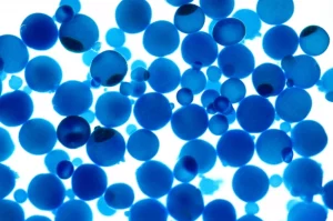 Fotografia macro ad alta definizione di microperle composite porose (alginato di calcio, gomma arabica, guscio d'uovo) immerse in una soluzione acquosa blu (blu di metilene), con alcune perle che mostrano il colorante adsorbito. Illuminazione controllata, obiettivo macro 100mm, alta definizione, messa a fuoco precisa sulla texture delle perle.