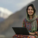 Fotografia stile reportage, una donna imprenditrice sorridente del Gilgit-Baltistan sui 35 anni, vestita con abiti tradizionali colorati ma moderni, utilizza un laptop seduta all'aperto con uno sfondo mozzafiato delle montagne del Karakorum, luce naturale del tardo pomeriggio, obiettivo 50mm f/1.8, profondità di campo ridotta per isolare la donna dallo sfondo ma mantenendo riconoscibile il paesaggio.