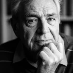 Fotografia ritratto di un uomo anziano (70 anni circa) dall'aria intellettuale e gentile, forse Martin Downer. Luce laterale morbida che evidenzia le linee del viso. Sfondo leggermente sfocato che potrebbe suggerire uno studio o una libreria. Stile bianco e nero filmico, obiettivo prime 35mm, profondità di campo media per mantenere il soggetto a fuoco ma ammorbidire lo sfondo.