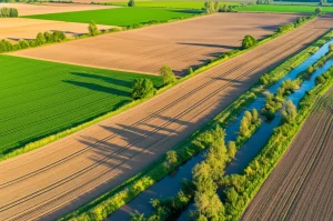 Paesaggio agricolo diversificato visto dall'alto, obiettivo grandangolare 10mm, che mostra campi coltivati intervallati da siepi, fasce tampone lungo un corso d'acqua e piccole aree naturali. Luce del primo mattino che crea lunghe ombre e accentua le texture. Lunga esposizione per rendere l'acqua del fiume liscia e setosa, simboleggiando l'integrazione tra agricoltura produttiva e protezione ambientale promossa dagli schemi agroambientali.