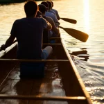 Primo piano di una canoa tradizionale Nativa Americana in legno, intagliata a mano, che scivola dolcemente su acque calme al tramonto. Si vedono le pagaie mosse ritmicamente da figure non ben definite all'interno. La luce calda del sole basso crea riflessi dorati sull'acqua e mette in risalto le venature del legno. Obiettivo zoom teleobiettivo 100mm, movimento leggermente sfocato per dare senso di avanzamento, atmosfera pacifica e comunitaria.