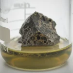 Immagine fotorealistica, obiettivo macro 90mm, di un campione di roccia ultramafica scura (dunite) parzialmente immerso in un liquido torbido (biolisciviante) in un becher da laboratorio, con dettagli precisi sulla superficie della roccia che mostra segni di erosione chimica, illuminazione controllata da laboratorio che enfatizza le texture.