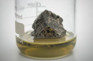 Immagine fotorealistica, obiettivo macro 90mm, di un campione di roccia ultramafica scura (dunite) parzialmente immerso in un liquido torbido (biolisciviante) in un becher da laboratorio, con dettagli precisi sulla superficie della roccia che mostra segni di erosione chimica, illuminazione controllata da laboratorio che enfatizza le texture.