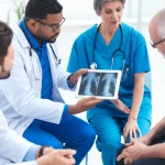 Fotografia realistica di un gruppo diversificato di professionisti sanitari e sociali (medico, infermiere, assistente sociale, fisioterapista) in cerchio, che discutono animatamente ma costruttivamente il caso di un paziente con artrite infiammatoria, le cui radiografie sono visibili su un tablet al centro. Ambiente clinico moderno e luminoso. Obiettivo prime 35mm, profondità di campo, luce naturale morbida.
