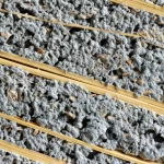 Primo piano macro, 100mm, di una sezione trasversale della malta NRG-WOOD. Si vedono chiaramente le fibre di legno riciclato, alcune con un aspetto leggermente ceroso (PCM), incapsulate da uno strato sottile e granuloso (resina e sabbia) e immerse nella matrice cementizia grigia. Illuminazione controllata laterale per enfatizzare le diverse texture e la composizione stratificata. High detail, precise focusing.