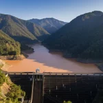 Vista aerea di una grande diga in una valle montuosa, con il serbatoio parzialmente riempito d'acqua torbida vicino all'ingresso del fiume dopo un evento piovoso intenso. Obiettivo grandangolare 18mm, messa a fuoco nitida, luce del tardo pomeriggio che crea ombre lunghe.