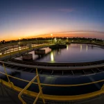 Fotografia grandangolare di un impianto di trattamento delle acque reflue al tramonto. Obiettivo 15mm wide-angle, lunga esposizione per rendere l'acqua nelle vasche liscia come seta, luci dell'impianto accese, cielo con colori caldi, messa a fuoco nitida sull'intera struttura, trasmettendo la scala del monitoraggio ambientale.