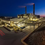 Impianto di termovalorizzazione moderno e pulito visto dall'alto al crepuscolo, con nastri trasportatori che movimentano rifiuti urbani verso l'area di combustione. Luci industriali accese, fotografia grandangolare 18mm, lunga esposizione per scie luminose, messa a fuoco nitida sull'intero impianto, concetto di waste-to-energy.