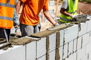 Immagine fotorealistica, obiettivo grandangolare 24mm, che mostra un cantiere edile sostenibile con blocchi geopolimerici realizzati in tufo vulcanico e scorie d'altoforno, con operai che applicano la malta, messa a fuoco nitida.