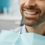 Ritratto di un paziente adulto sorridente che indossa allineatori trasparenti con attacchi per elastici di Classe II visibili sul canino superiore, sfondo sfocato di uno studio dentistico moderno e luminoso, obiettivo prime 35mm, profondità di campo ridotta per mettere a fuoco il sorriso.