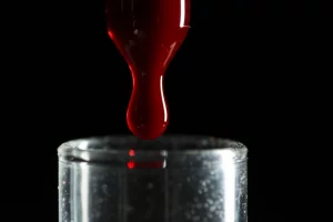 Immagine fotorealistica di una goccia di sangue che cade in una provetta, macro lens 100mm, illuminazione drammatica controllata, alta definizione, sfondo scuro.