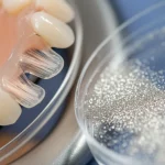 Immagine fotorealistica, obiettivo macro 80mm. Un ponte dentale provvisorio in PMMA traslucido rinforzato con microfibre di vetro visibili, appoggiato su un vassoio dentale sterile. Accanto, una piccola piastra di Petri contenente polvere di nanoparticelle d'argento scintillanti. Alto dettaglio, messa a fuoco precisa sulle texture, illuminazione controllata da studio dentistico, sfondo leggermente sfocato.