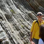 Turisti interessati osservano da vicino una spettacolare formazione rocciosa stratificata in un geoparco. Fotografia di reportage, obiettivo zoom 35mm, profondità di campo media che mantiene a fuoco sia le persone che la roccia, luce naturale brillante, espressioni di curiosità e meraviglia sui volti.