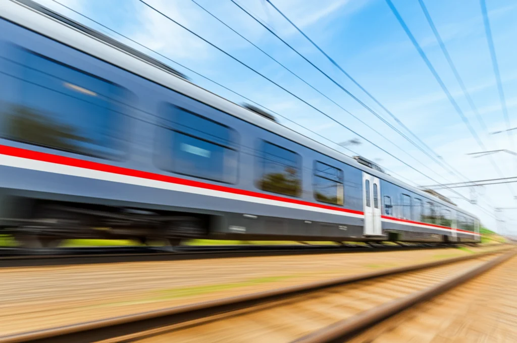 Vista dinamica di un treno passeggeri che sfreccia su un binario, catturata con un teleobiettivo zoom 100-400mm. Scatto ad alta velocità per congelare il movimento del treno ma con leggero motion blur sui binari sottostanti, luce solare diretta che evidenzia i dettagli del treno e del paesaggio circostante, tracciamento del movimento attivo.