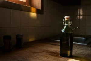 Bottiglia di olio extra vergine d'oliva Cobrançosa in vetro ambrato semiaperta su un piano di cucina in legno rustico. Metà della scena è illuminata da una luce solare calda che entra da una finestra, l'altra metà è in ombra profonda, simboleggiando le diverse condizioni di conservazione domestica. Obiettivo 50mm, profondità di campo media, luce naturale controllata per un effetto realistico e suggestivo.