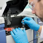 Immagine photorealistica di un tecnico di laboratorio che indossa occhiali protettivi e guanti mentre carica con attenzione una fiala (vial) in un campionatore automatico (autosampler) di un gascromatografo (GC/FID). Sullo sfondo, strumentazione da laboratorio moderna e pulita. Profondità di campo ridotta per mettere a fuoco l'azione. Obiettivo prime 35mm, illuminazione da laboratorio controllata.