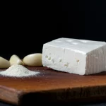 Fotografia di cibo still life: un pezzo invitante di formaggio morbido bianco su un tagliere scuro, con accanto spicchi d'aglio e un piccolo mucchietto di polvere fine (l'estratto incapsulato). Luce laterale drammatica, obiettivo macro 90mm, alta definizione, focus selettivo sulla texture del formaggio.