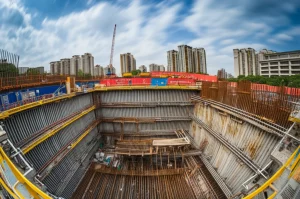 Immagine panoramica grandangolare di un cantiere di scavo profondo a Ningbo, Cina, in un'area urbana densamente popolata, con muri di diaframma in cemento armato e sistemi di supporto in acciaio visibili sotto un cielo parzialmente nuvoloso, obiettivo grandangolare 12mm, lunga esposizione per sfumare le nuvole, messa a fuoco nitida sull'intera scena.