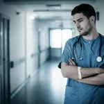 Primo piano di un infermiere pensieroso in un corridoio d'ospedale, luce soffusa laterale, obiettivo 35mm, profondità di campo ridotta, toni blu e grigi duotone, che riflette sulle sfide della rimozione precoce del catetere urinario post-chirurgia spinale.