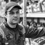 Ritratto ambientato di un lavoratore 'colletto blu' (uomo o donna, età media) in un ambiente di vendita al dettaglio o magazzino, sguardo pensieroso o stanco. Obiettivo 35mm, profondità di campo ridotta per sfocare lo sfondo, luce naturale laterale, bianco e nero.