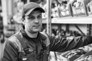 Ritratto ambientato di un lavoratore 'colletto blu' (uomo o donna, età media) in un ambiente di vendita al dettaglio o magazzino, sguardo pensieroso o stanco. Obiettivo 35mm, profondità di campo ridotta per sfocare lo sfondo, luce naturale laterale, bianco e nero.