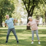 Immagine fotorealistica di un uomo anziano e una donna anziana che fanno esercizi leggeri di stretching per la schiena in un parco luminoso, obiettivo 50mm, luce naturale del mattino, profondità di campo media per includere l'ambiente sereno.
