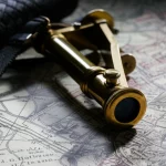 Primo piano del telemetro J.G. Hofmann appoggiato su una vecchia mappa militare del XIX secolo, con luce laterale drammatica che evidenzia i dettagli in ottone lucido, la copertura intrecciata scura e l'oculare. Obiettivo macro 100mm, alta definizione, messa a fuoco precisa sui dettagli incisi.