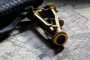 Primo piano del telemetro J.G. Hofmann appoggiato su una vecchia mappa militare del XIX secolo, con luce laterale drammatica che evidenzia i dettagli in ottone lucido, la copertura intrecciata scura e l'oculare. Obiettivo macro 100mm, alta definizione, messa a fuoco precisa sui dettagli incisi.