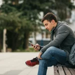 Ritratto di uno studente universitario seduto su una panchina del campus, che guarda pensieroso il suo smartphone ma con scarpe da corsa ai piedi e uno zaino sportivo accanto, profondità di campo ridotta per sfocare lo sfondo, obiettivo prime 35mm, luce naturale pomeridiana, che simboleggia la scelta tra dipendenza da smartphone e benefici dell'attività fisica.
