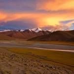 Paesaggio mozzafiato dell'altopiano tibetano al tramonto, obiettivo grandangolare 20mm, messa a fuoco nitida sulle montagne in lontananza e su un fiume serpeggiante, colori caldi, suggerendo la bellezza naturale ma anche la vulnerabilità ambientale legata all'inquinamento del suolo.