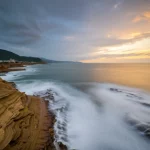 Paesaggio grandangolare della costa centrale di Taiwan al tramonto, con una rappresentazione grafica stilizzata che mostra le formazioni geologiche sottomarine e terrestri adatte allo stoccaggio di CO2. Wide-angle, 10mm, long exposure, smooth water.