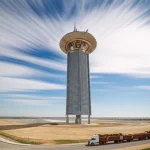 Fotografia paesaggistica realistica, obiettivo grandangolare 20mm, messa a fuoco nitida, lunga esposizione con nuvole effetto seta. Raffigura una futuristica torre solare a concentrazione integrata con un impianto di trattamento biomasse sotto un sole splendente, con camion che trasportano balle di biomassa visibili sulle strade circostanti, simboleggiando l'intersezione tra energia solare, biomassa e logistica per la produzione di idrogeno negli Stati Uniti.