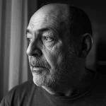 Ritratto intenso di un uomo di mezza età, sopravvissuto a tumore cerebrale, che guarda fuori da una finestra con espressione preoccupata ma resiliente, simboleggiando la lotta contro la tossicità finanziaria; fotografia 50mm prime lens, profondità di campo media, luce naturale laterale che crea contrasto, bianco e nero cinematografico.