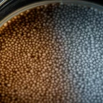 Immagine fotorealistica di granuli del composito feldspato/MnO2 che purificano acqua visibilmente contaminata da metalli pesanti in un becher di vetro da laboratorio, con una chiara distinzione tra acqua sporca e pulita, obiettivo macro 90mm, illuminazione drammatica laterale, alta definizione, messa a fuoco precisa sui granuli.