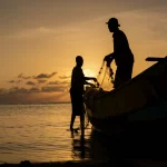 Immagine fotorealistica di un pescatore keniota sulla sua piccola barca al largo della costa, mentre tira su una rete al tramonto. Luce dorata, silhouette contro il sole basso sull'orizzonte marino. Obiettivo zoom 50mm, profondità di campo media, stile evocativo e pieno di speranza.