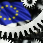 Primo piano di ingranaggi metallici complessi che si incastrano, con una bandiera dell'UE leggermente sfocata sullo sfondo, fotografia macro 100mm, illuminazione controllata per evidenziare i dettagli metallici, simboleggia la complessa macchina tecnocratica della governance economica UE e le sue radici neoliberiste.