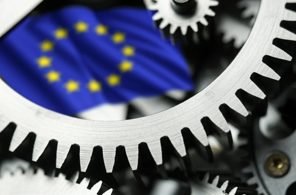 Primo piano di ingranaggi metallici complessi che si incastrano, con una bandiera dell'UE leggermente sfocata sullo sfondo, fotografia macro 100mm, illuminazione controllata per evidenziare i dettagli metallici, simboleggia la complessa macchina tecnocratica della governance economica UE e le sue radici neoliberiste.