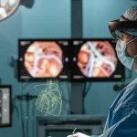 Immagine fotorealistica di un chirurgo in sala operatoria che indossa un visore HoloLens2, osservando un ologramma 3D fluttuante dell'anatomia vascolare del paziente accanto a un monitor laparoscopico. Illuminazione chirurgica intensa, messa a fuoco precisa sull'interazione tra chirurgo e tecnologia, sfondo leggermente sfocato per profondità di campo, lente prime 50mm.