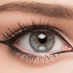 Macro fotografia di un occhio femminile truccato con eyeliner nero applicato sulla rima esterna, con alcune micro-particelle visibili vicino alla linea delle ciglia, suggerendo potenziale irritazione. Obiettivo macro 100mm, alta definizione, illuminazione laterale per enfatizzare la texture della pelle e del trucco.