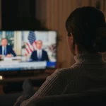Immagine fotorealistica stile reportage, obiettivo 35mm, che mostra una persona vista di spalle guardare contemporaneamente due schermi: uno con una chat video con familiari in un paese lontano, l'altro con un notiziario politico americano. Luce soffusa, profondità di campo che sfoca leggermente lo sfondo della stanza negli USA, simboleggiando il legame tra paese d'origine e residenza.